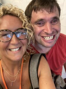 Two people smiling into the camera taking a selfie. One is wearing glasses and has blond curly hair. The other is wearing a red t-shirt and has short brown hair. 